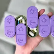 3D-printed weekly AM/PM pill organizer in purple and green, with sliding lids for easy access, held in hand for size reference.