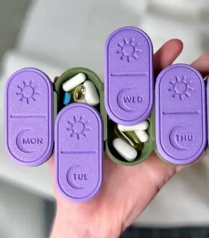 3D-printed weekly AM/PM pill organizer in purple and green, with sliding lids for easy access, held in hand for size reference.